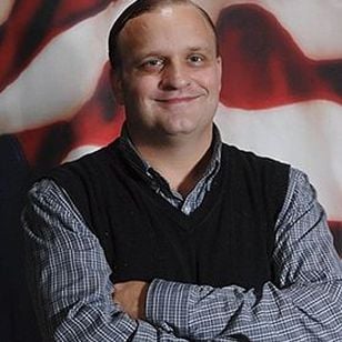 Jim Donovan smiling with his arms crossed, wearing a black sweater vest over a checked shirt.