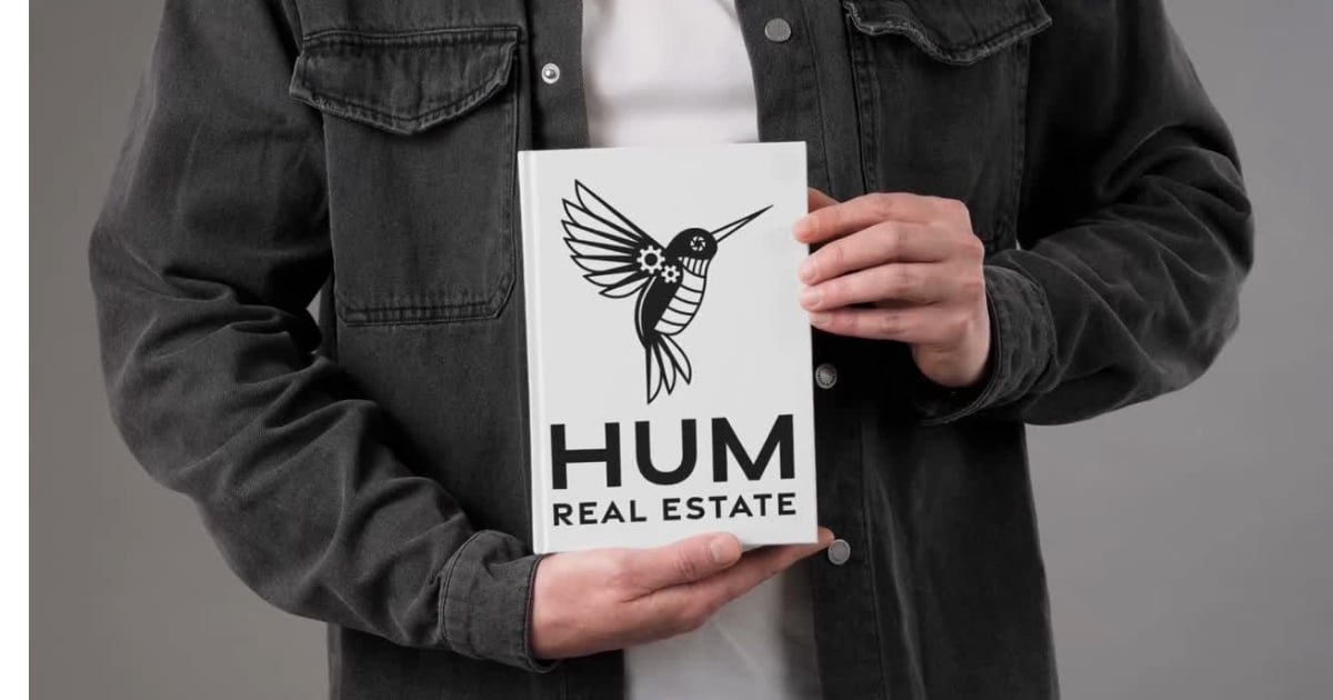 Person in a dark denim jacket holding a HUM Real Estate sign with a hummingbird emblem.