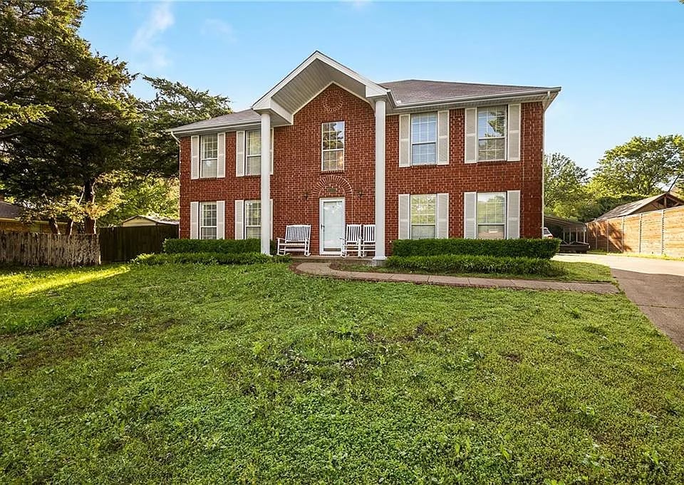 Two-story red brick house with white-trimmed windows, front lawn, and chairs by the entry.