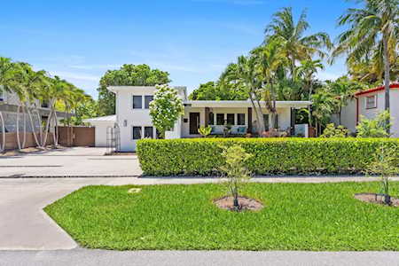 Modern white home behind a clipped hedge, with a green lawn, palms, and a paved driveway.