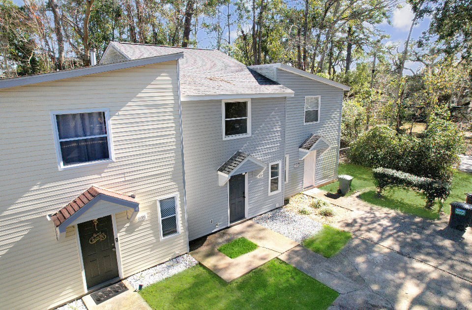 Two connected townhouse units with light gray siding, small porches, and manicured lawns are surrounded by trees and a sunny sky. A peaceful suburban setting.