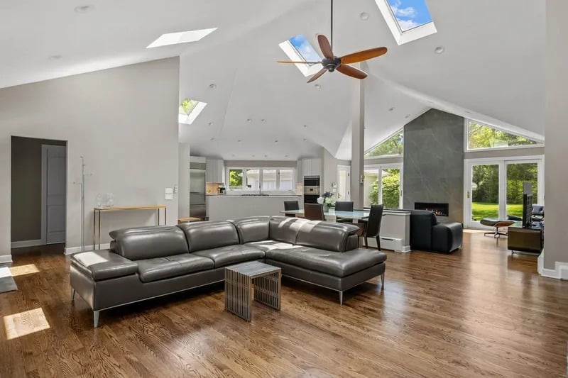 Open-plan living room with a gray sectional, vaulted skylit ceiling, stone fireplace, and windows overlooking a lawn.