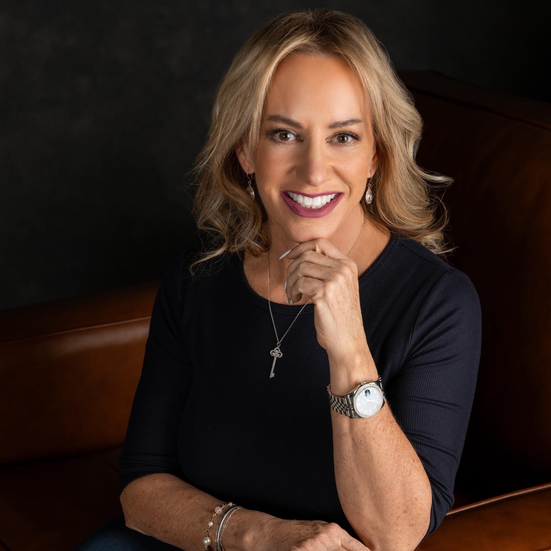 Linda Traurig smiles in a black top, wearing a key pendant necklace and silver wristwatch.