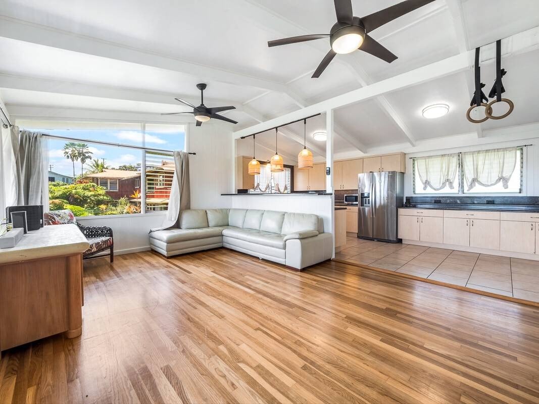 Open-plan living room and kitchen with a white sectional, wood floors, and large windows overlooking greenery.