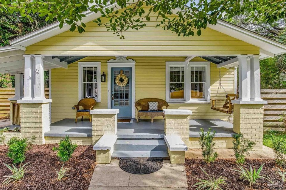 Pale yellow cottage with a broad covered porch, white columns, and a hanging swing.
