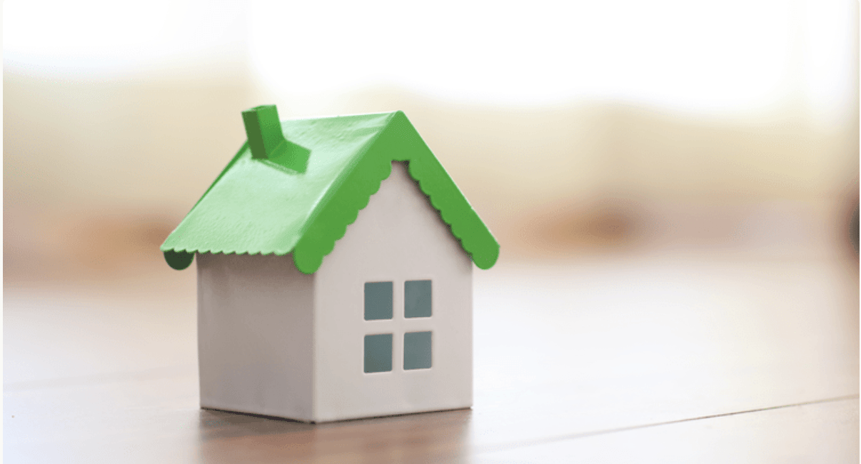 A small white model house with a bright green roof sits on a tabletop.
