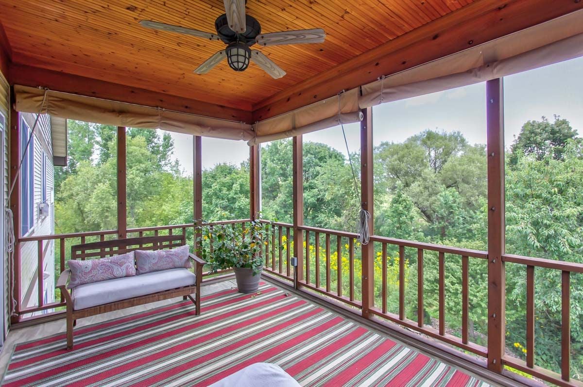 Screened porch with a wood ceiling, cushioned bench, striped rug, and leafy tree views.