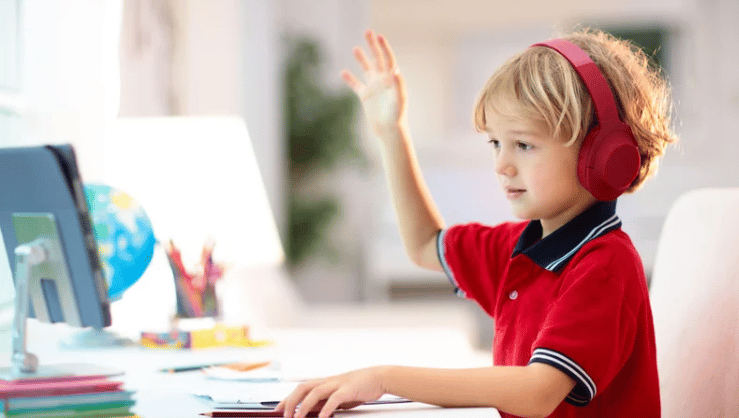 Child raising a hand at a desk while wearing red headphones.
