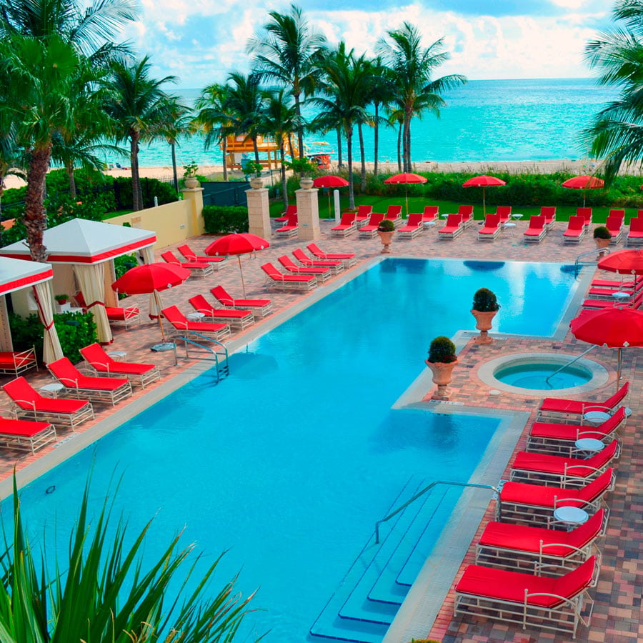 Outdoor resort pool with a circular spa, red loungers, palm trees and ocean views.