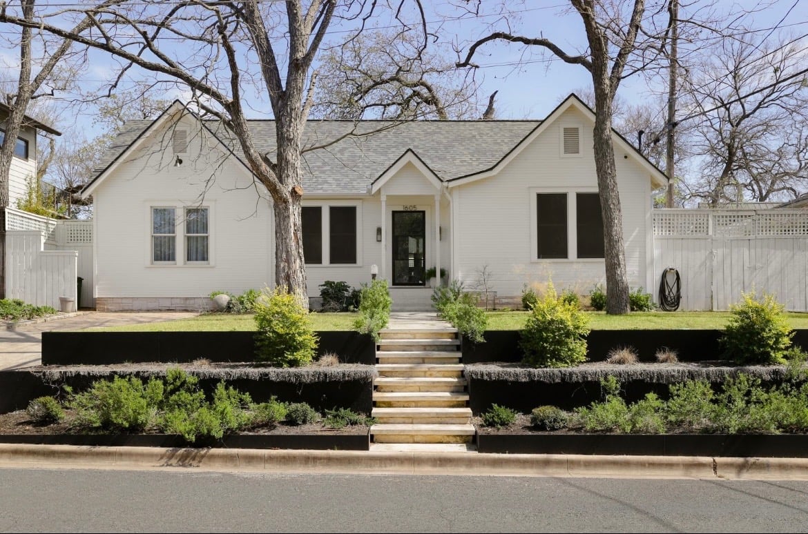 White single-story home with a central entry, broad front steps, tiered landscaping, and mature trees.