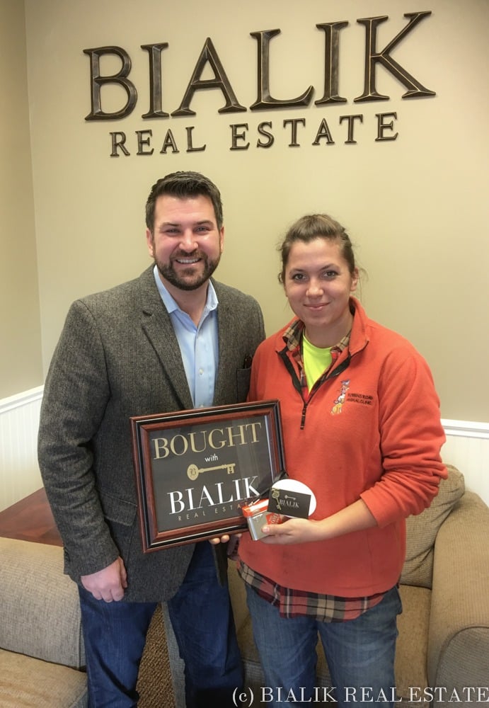 A man and woman pose with a framed “Bought with Bialik” sign beneath the office wall sign.