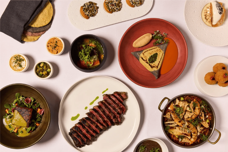Restaurant spread with sliced grilled steak, colorful small plates, salads, and a bowl of roasted ingredients.