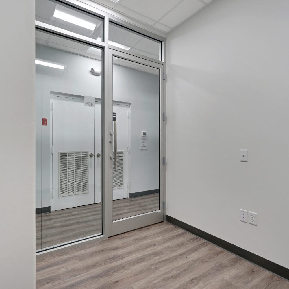 Empty office with a glass partition and door, gray wood-look flooring, and white walls.