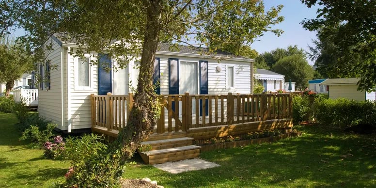 White mobile home with blue shutters and a raised wooden deck beneath mature trees.