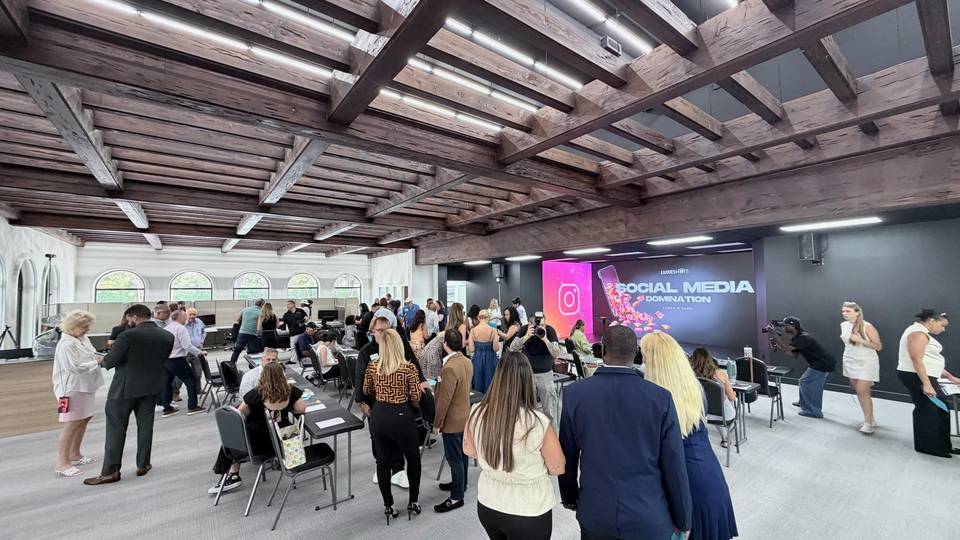 Attendees gather at tables in a large room with wooden ceiling beams, facing a screen that reads Luxeshots Social Media Domination