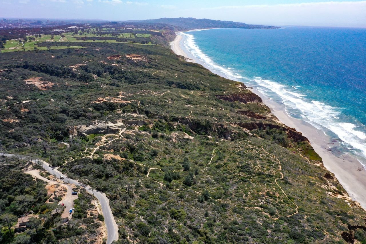 Aerial view of a rugged coastline, sandy beach, and blue ocean beside green coastal hills.