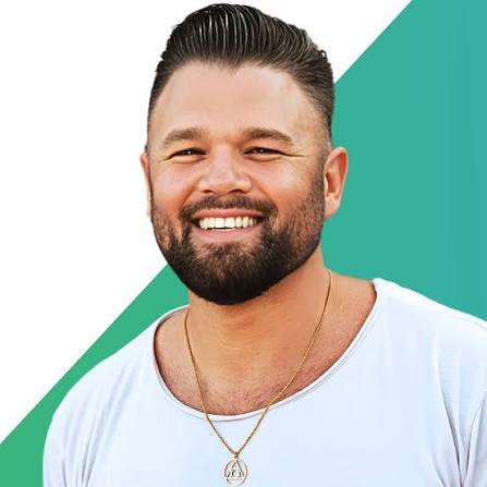 Smiling bearded man in a white shirt and gold pendant necklace against a teal geometric background.