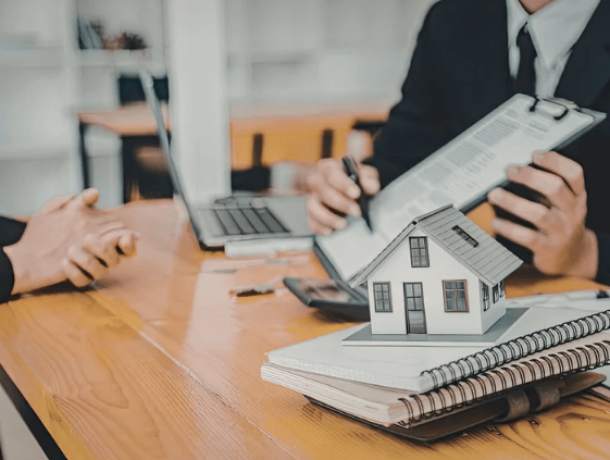 People review real estate documents beside a miniature house model and laptop.