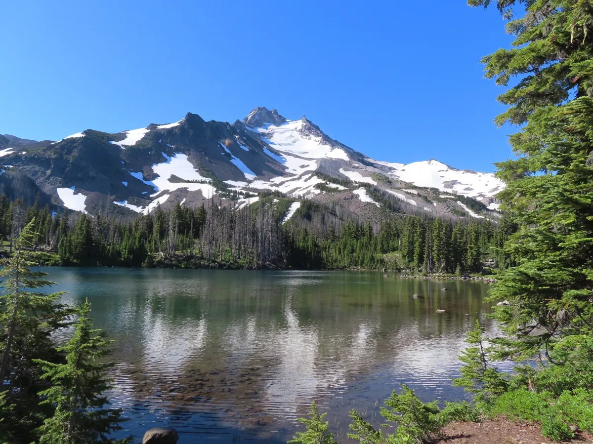 Mountain lake beneath a snow-covered peak, framed by evergreen forest.