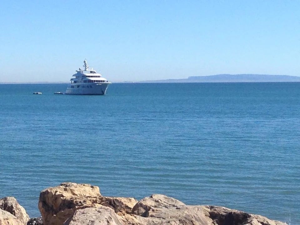 White yacht offshore on calm blue water, seen from a rocky shoreline.