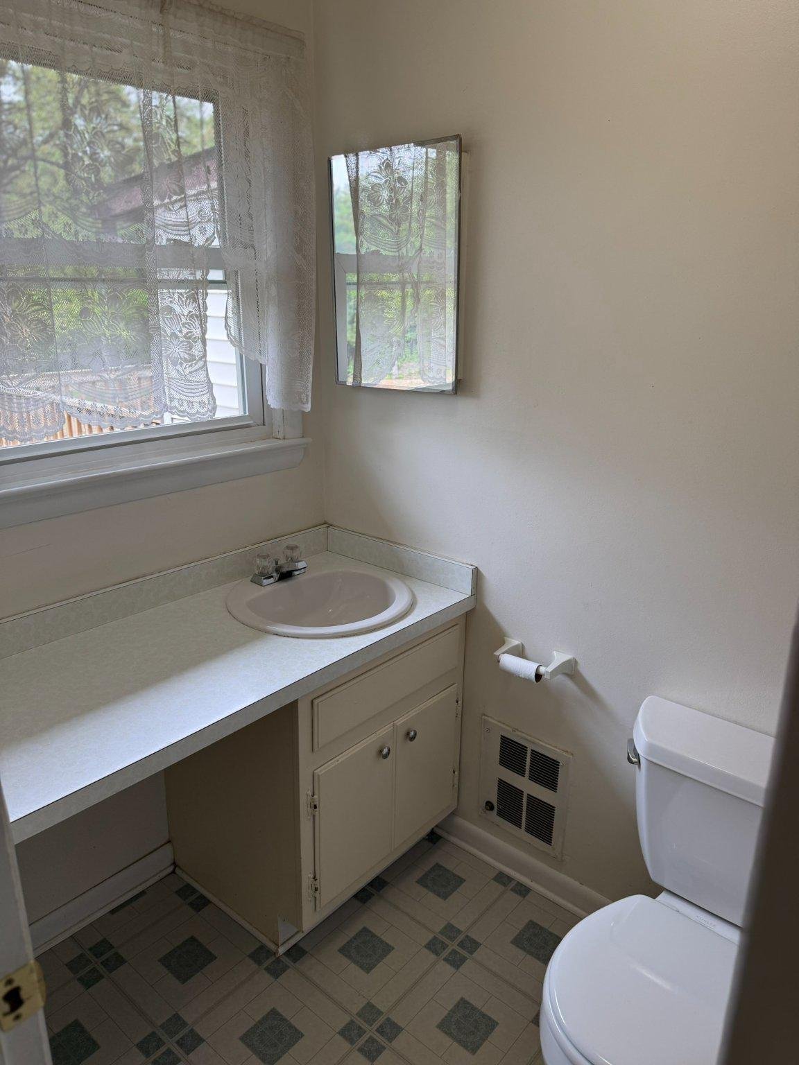 Bathroom with a white vanity sink, toilet, lace-curtained window, and patterned tile floor.