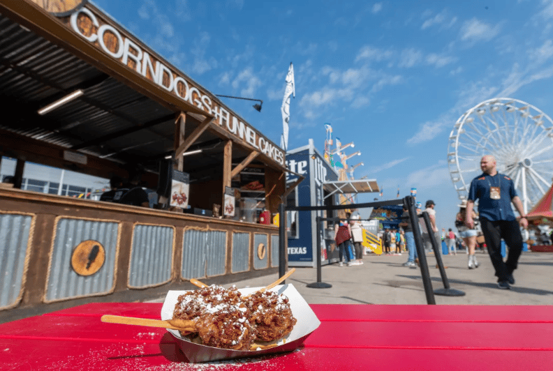 Powdered-sugar-covered fried treat on a red table at a rodeo food stand, with a Ferris wheel behind it.