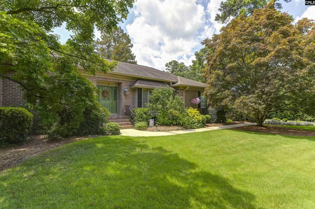Brick home with a green front door, landscaped lawn, mature trees, and an American flag.