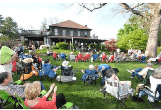 People gather in lawn chairs on a grassy lawn in front of a two-story house.