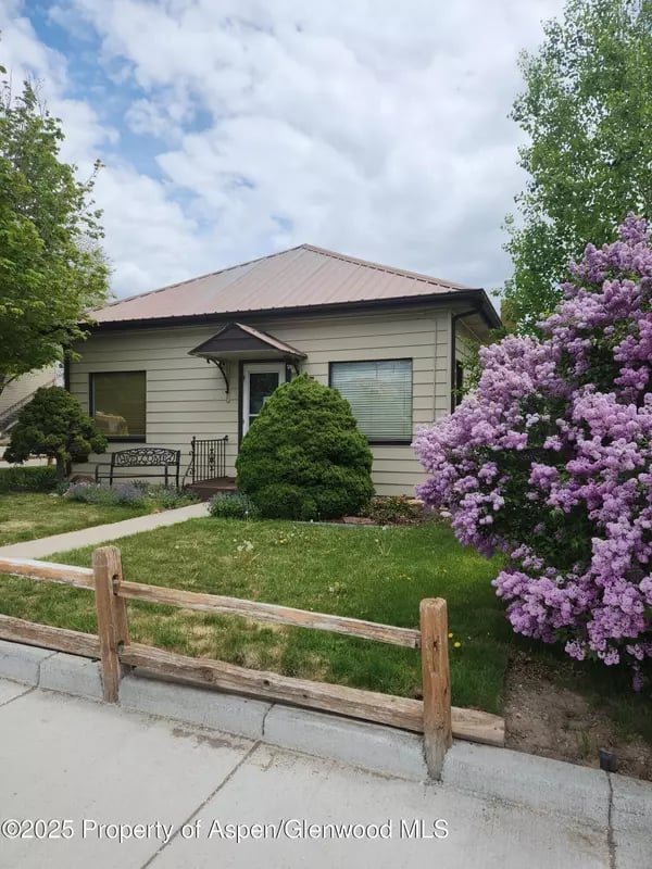 Single-story home with a metal roof, front lawn, wooden fence, and blooming lilac bush.