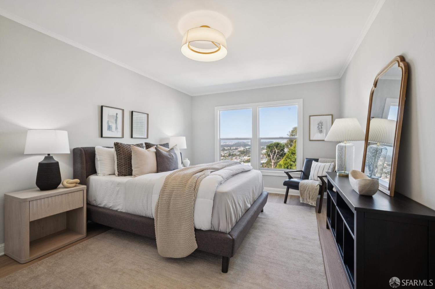 Staged bedroom with neutral bedding, a tall mirror, and a window overlooking trees and the city.