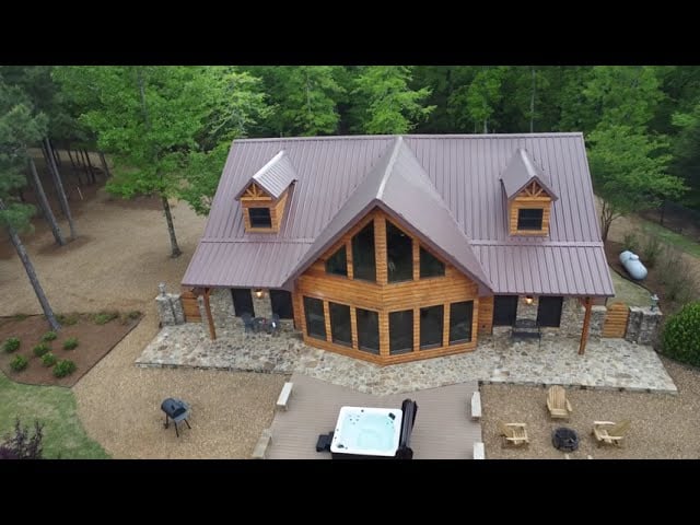 Wood-and-stone cabin with two roof dormers, a broad patio, and a hot tub, surrounded by trees.