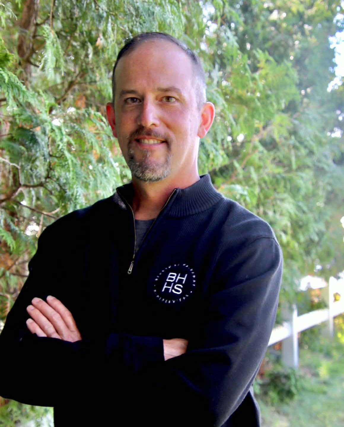 Eric Bottorff stands with folded arms in a navy sweater among leafy trees.