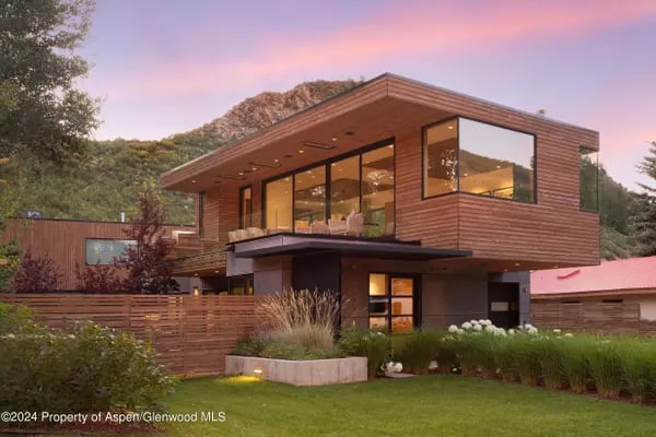 Modern wood-sided home with large windows and landscaped lawn at dusk, backed by hills.