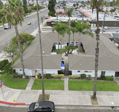 Aerial view of low-rise buildings with gray roofs, palm trees, and lawns beside a street.