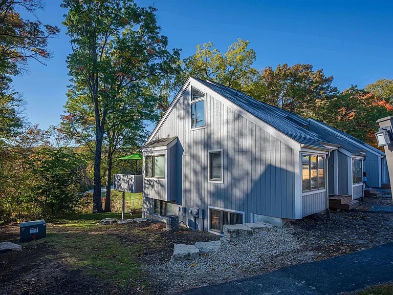Gray-sided home with a steep gabled roof beside a gravel drive and autumn trees.