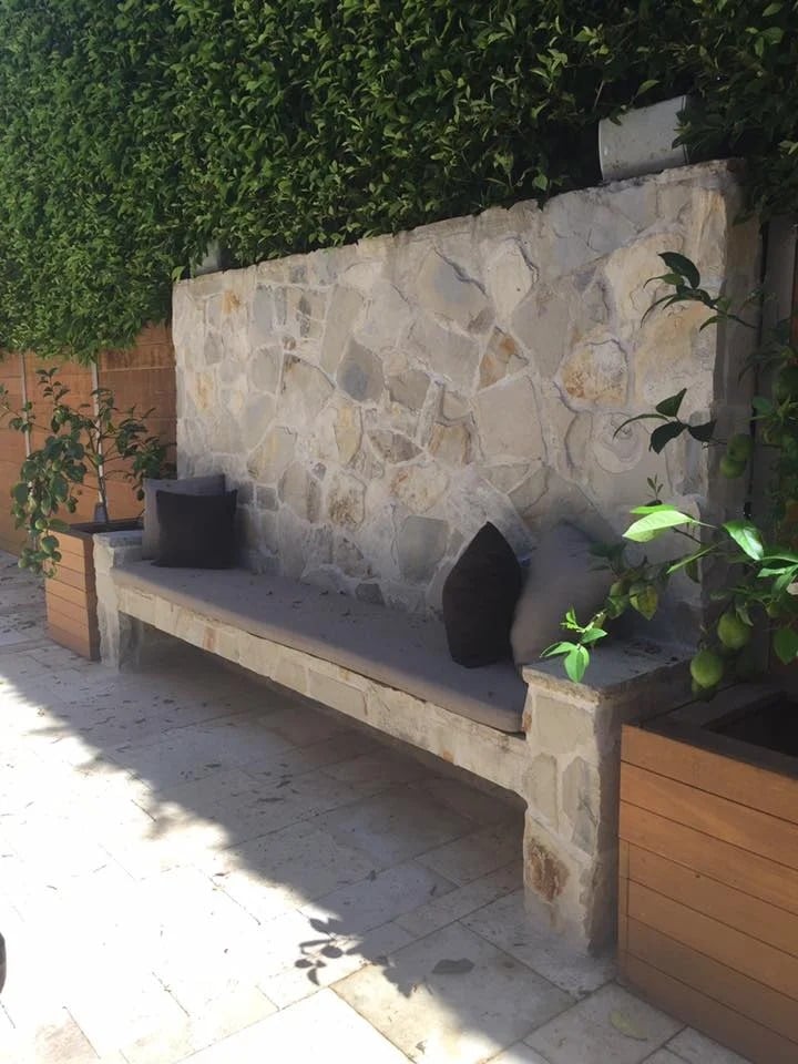 Built-in stone patio bench with dark cushions beneath a leafy hedge.