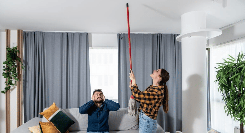 A man covers his ears on a sofa as a woman reaches toward the ceiling with a mop.