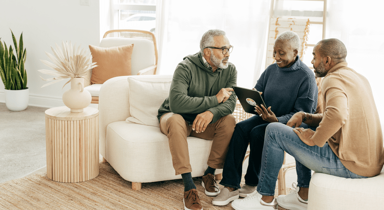 Three people sit on a white couch in a bright room, discussing something on a tablet. They seem engaged and happy. A vase and plant are in the background.