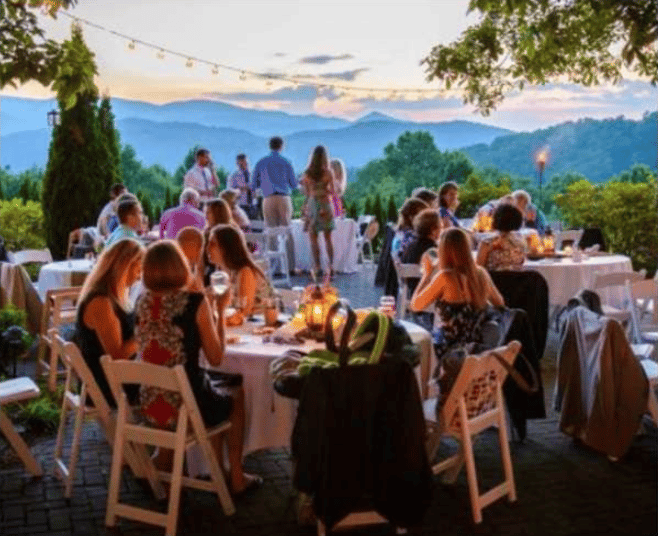 Guests dine outdoors beneath string lights with layered mountain views at sunset.