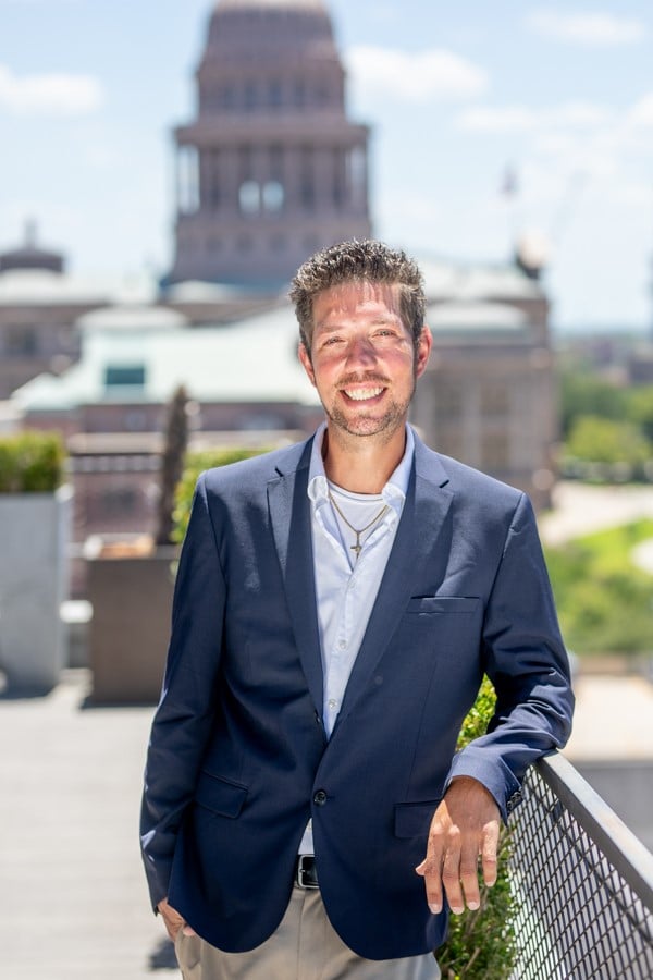 Chris Hitchcock smiles in a navy suit beside a railing, with a domed building behind him.