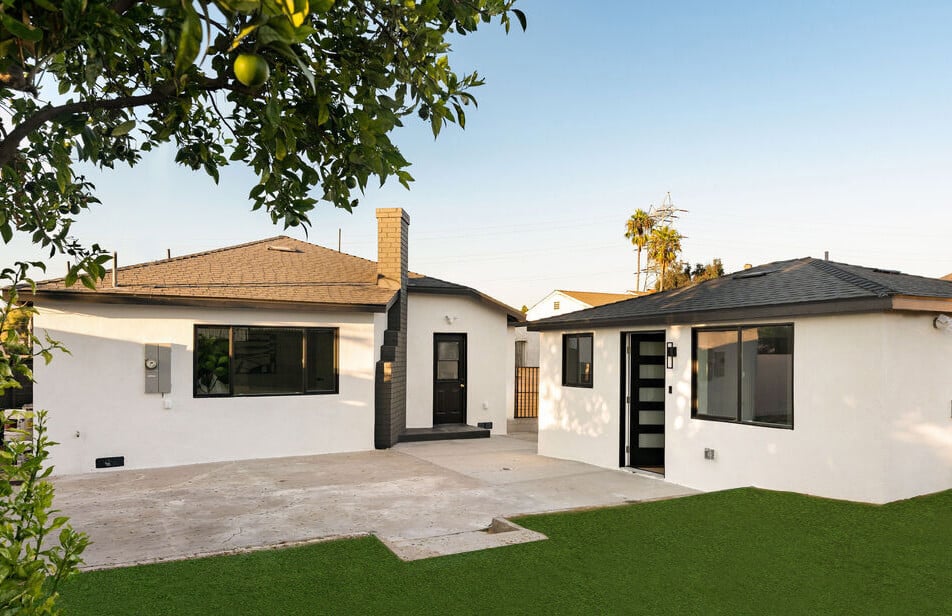 Two white single-story homes face a paved courtyard beside a small lawn and citrus tree.