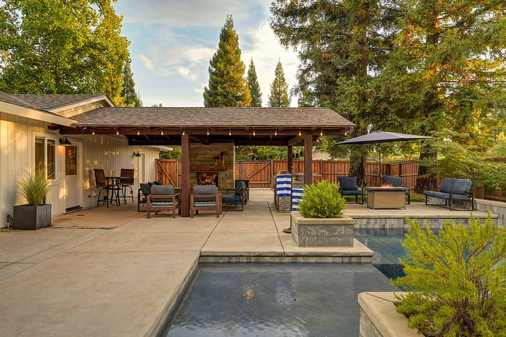 Backyard pool beside a covered patio with outdoor seating and a stone fireplace.