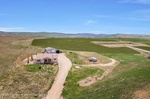 Rural home and outbuildings beside a winding dirt driveway, surrounded by open grassy land.