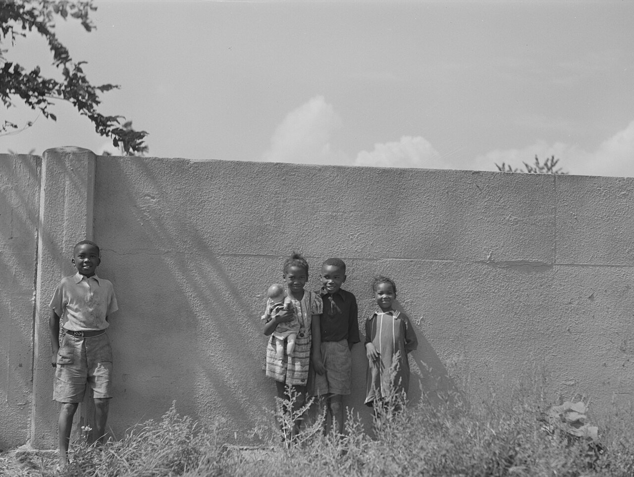 Five children stand beside a tall wall; one holds an infant, with weeds in the foreground.