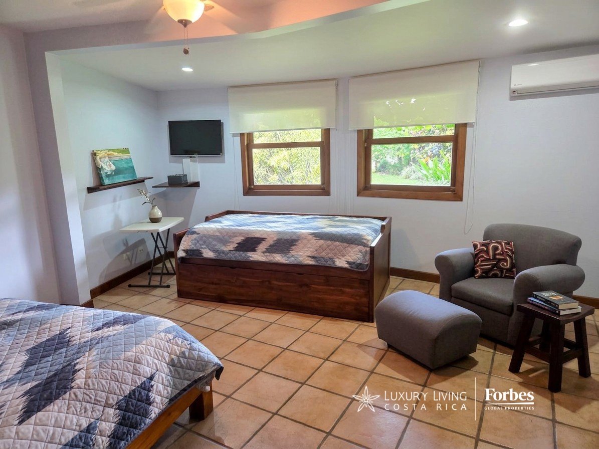 Furnished bedroom with two wooden beds, tiled floors, and windows overlooking greenery.