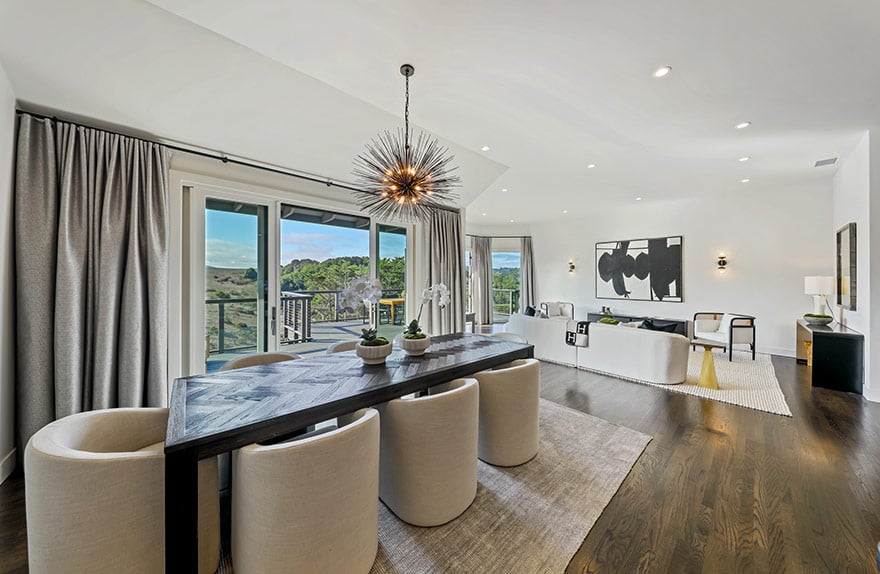 Open-plan dining and living room with cream chairs, a sculptural chandelier, and glass doors overlooking green hills.