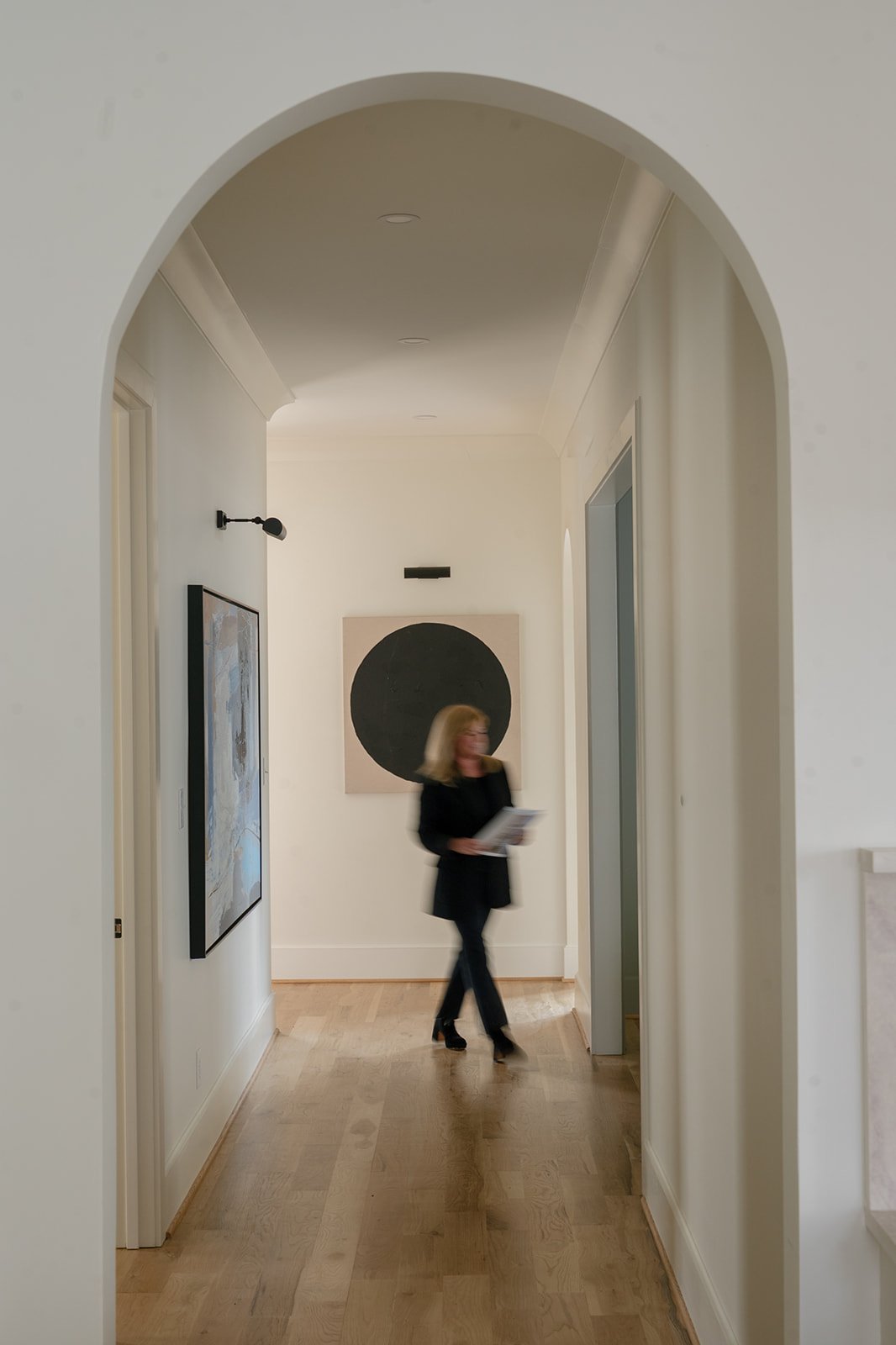 Woman carrying papers walks through a modern hallway with pale wood floors and an arched doorway.