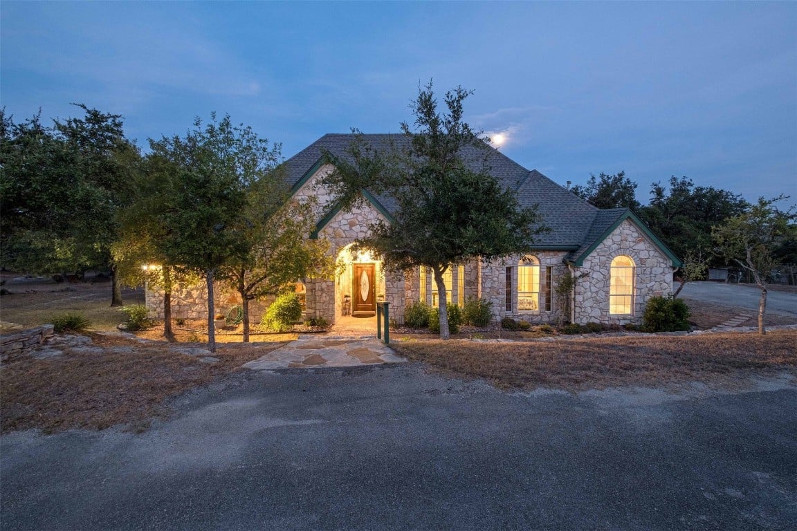 Stone-front home with a steep roof and glowing windows at dusk, framed by mature trees.