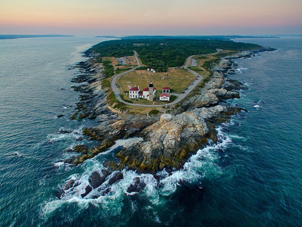 Aerial view of a lighthouse on a rocky peninsula, with ocean waves and a winding coastal road.
