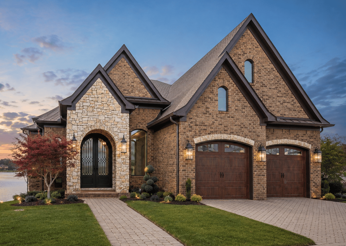 Brick and stone lakeside home with steep gables, arched entry, and two wooden garage doors at dusk.